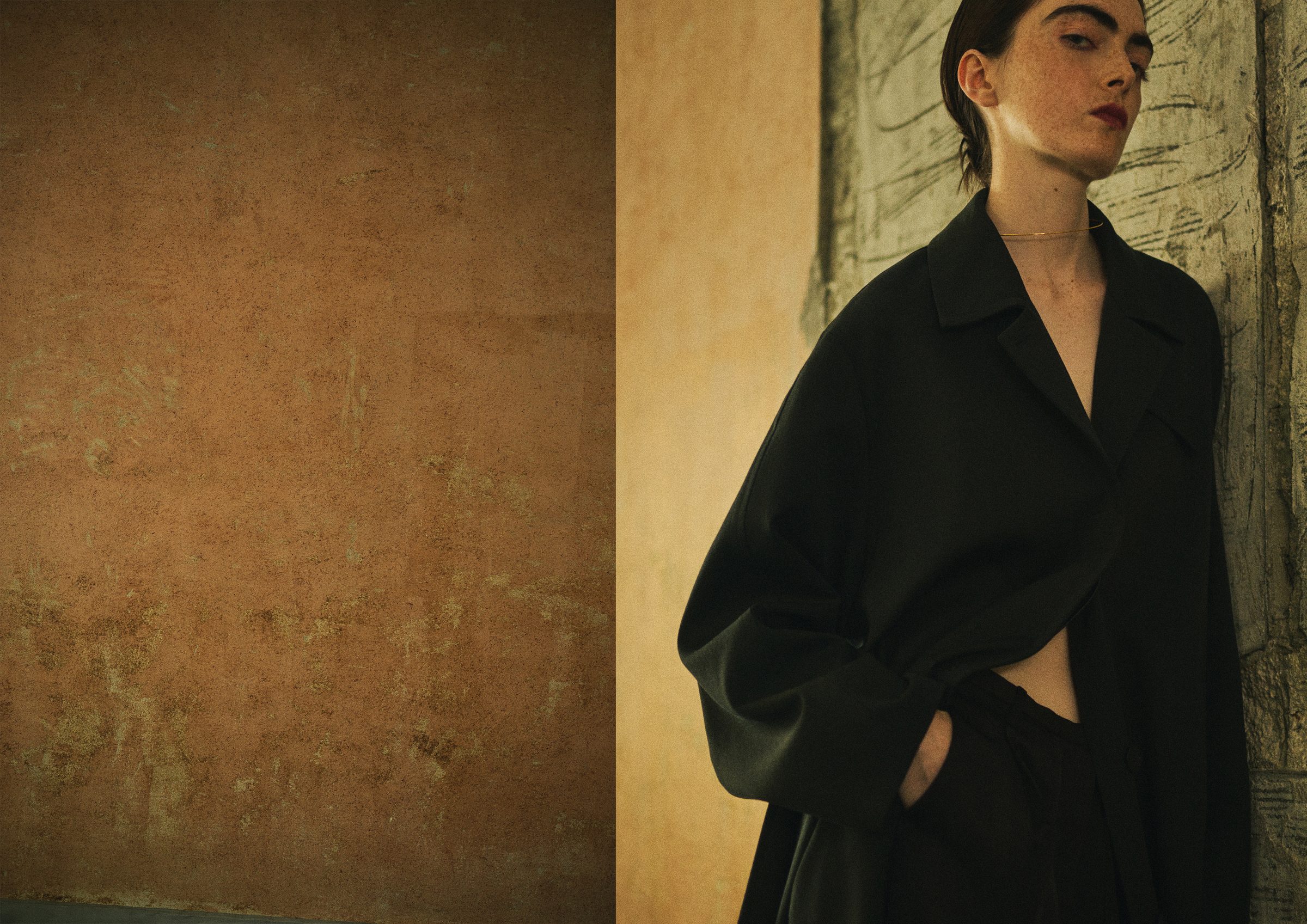 A model in a long black coat against a plaster wall