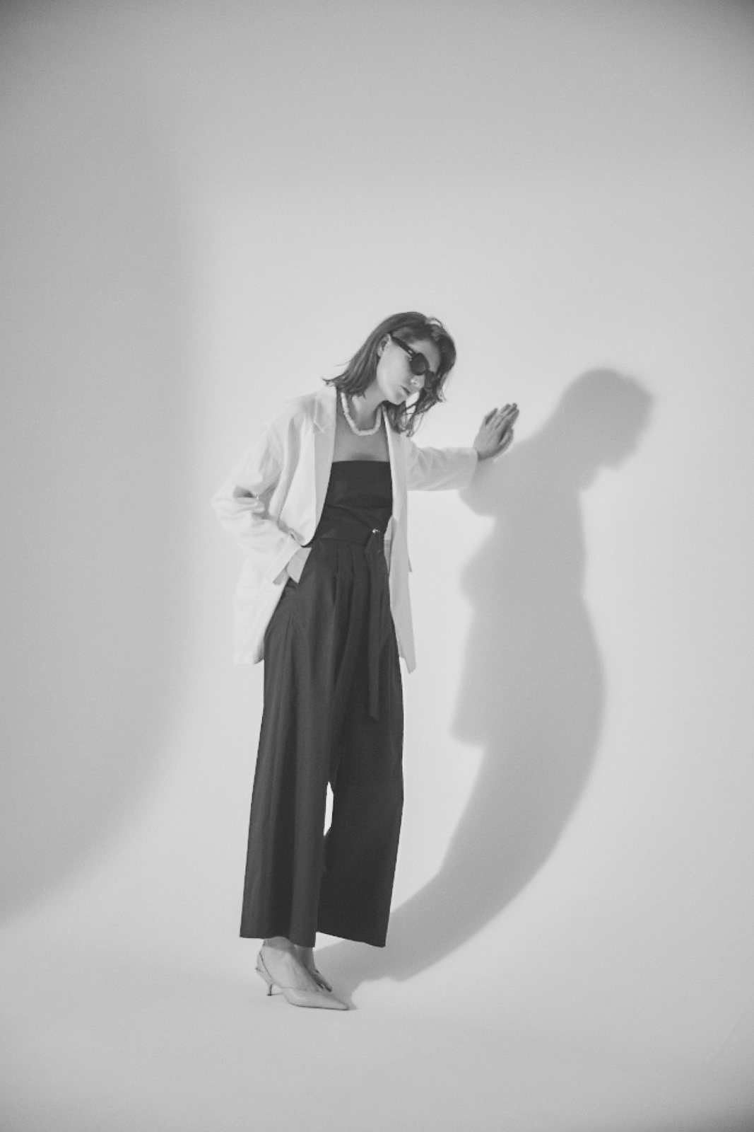 Black-and-white portrait of a model in a white jacket and black wide trousers leaning on a wall