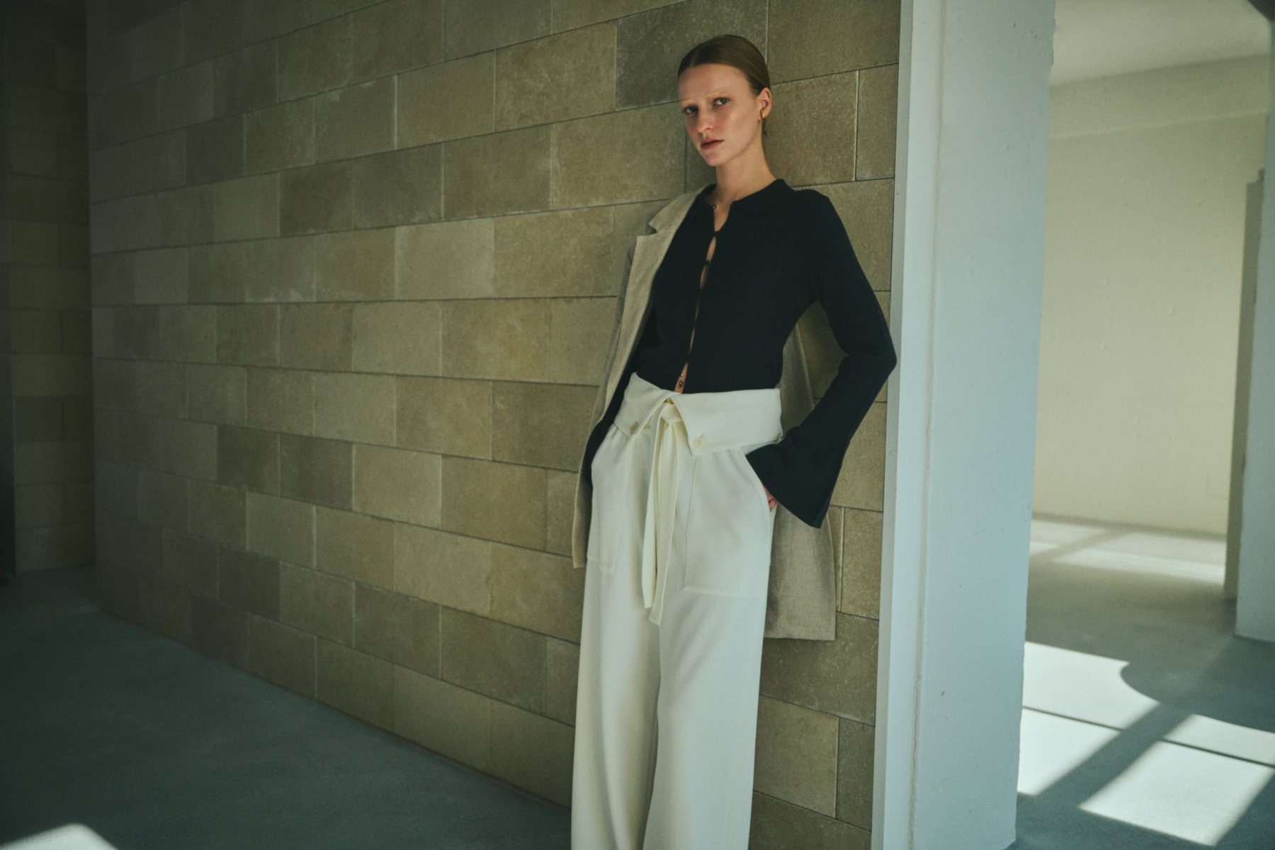 Model in a black cardigan and cream trousers against a stone wall